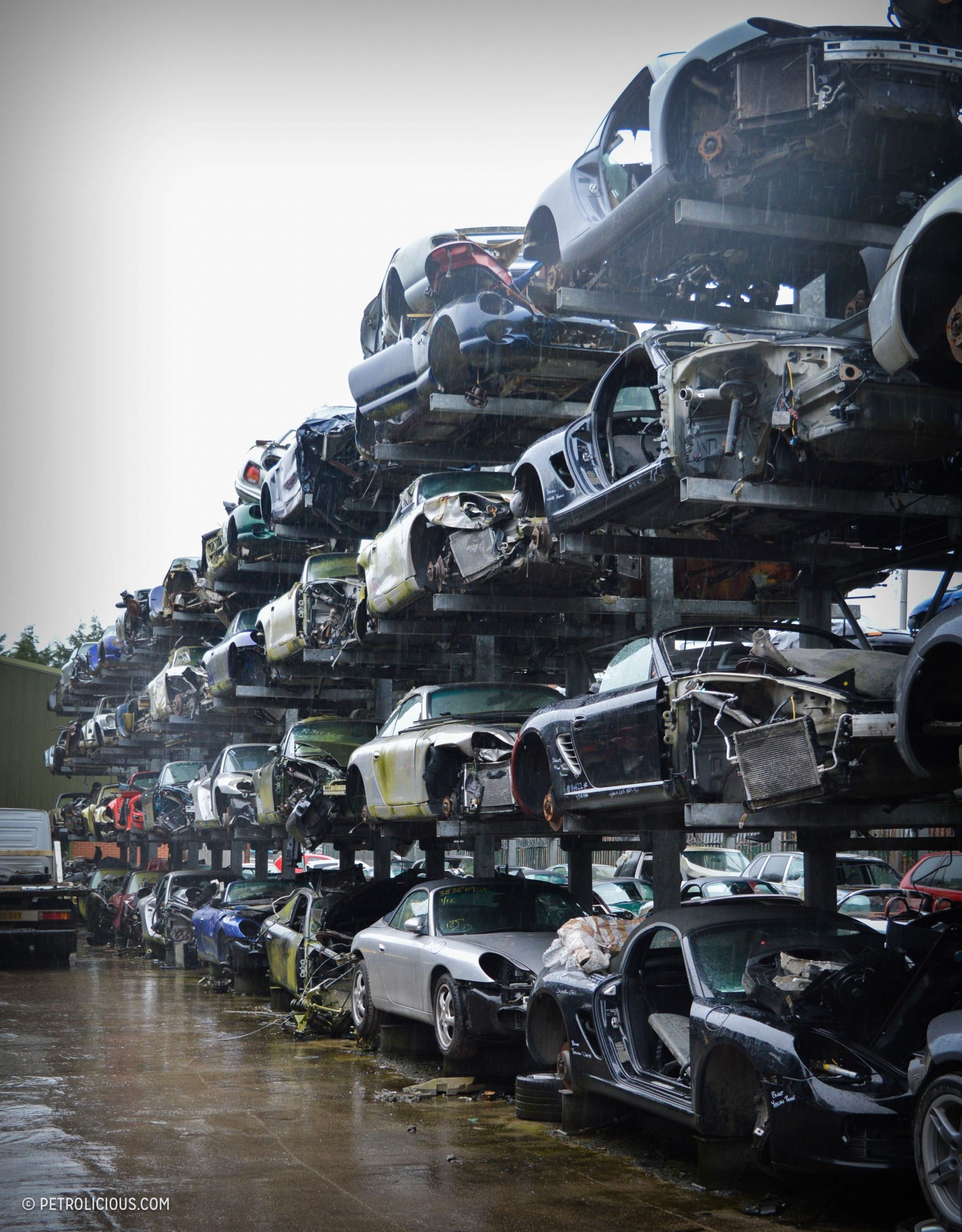 Vertical racks filled with luxury cars at a UK car scrap yard, showcasing Porsches and other high-end vehicles.